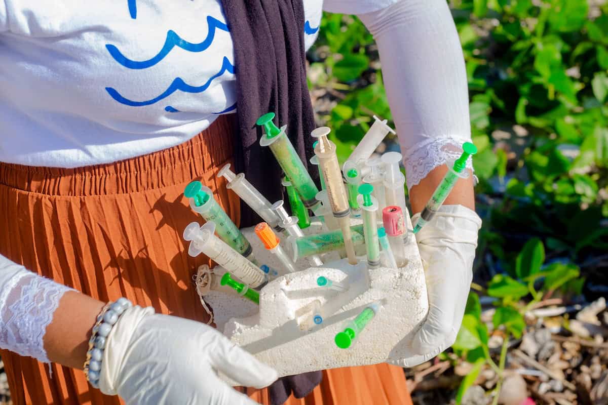 A person wearing gloves holds a block of styrofoam pierced with numerous discarded medical syringes collected during a beach cleanup. The individual wears a white shirt with a wave design and a rust-colored skirt.
