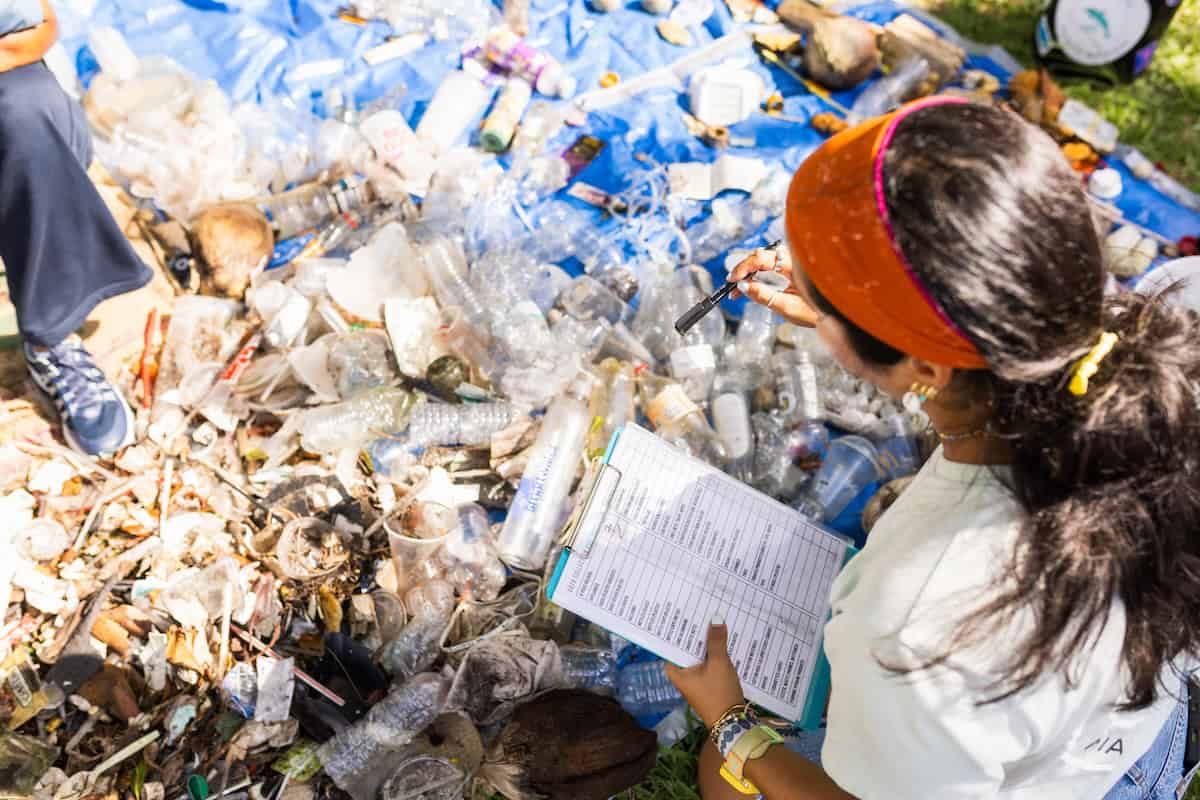 A woman wearing an orange headband and white shirt records data on a clipboard while sorting through a large pile of plastic waste and debris on a blue tarp during a coastal cleanup.