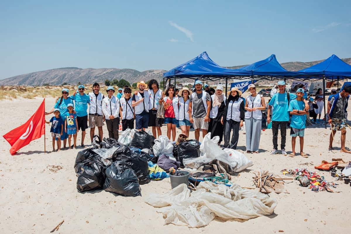 A large group of volunteers from the TunSea Organization stands on a sandy beach in Tunisia, smiling in front of piles of collected trash, including plastic bags, nets, and metal cans, after a cleanup event. Blue tents and mountains are visible in the background.