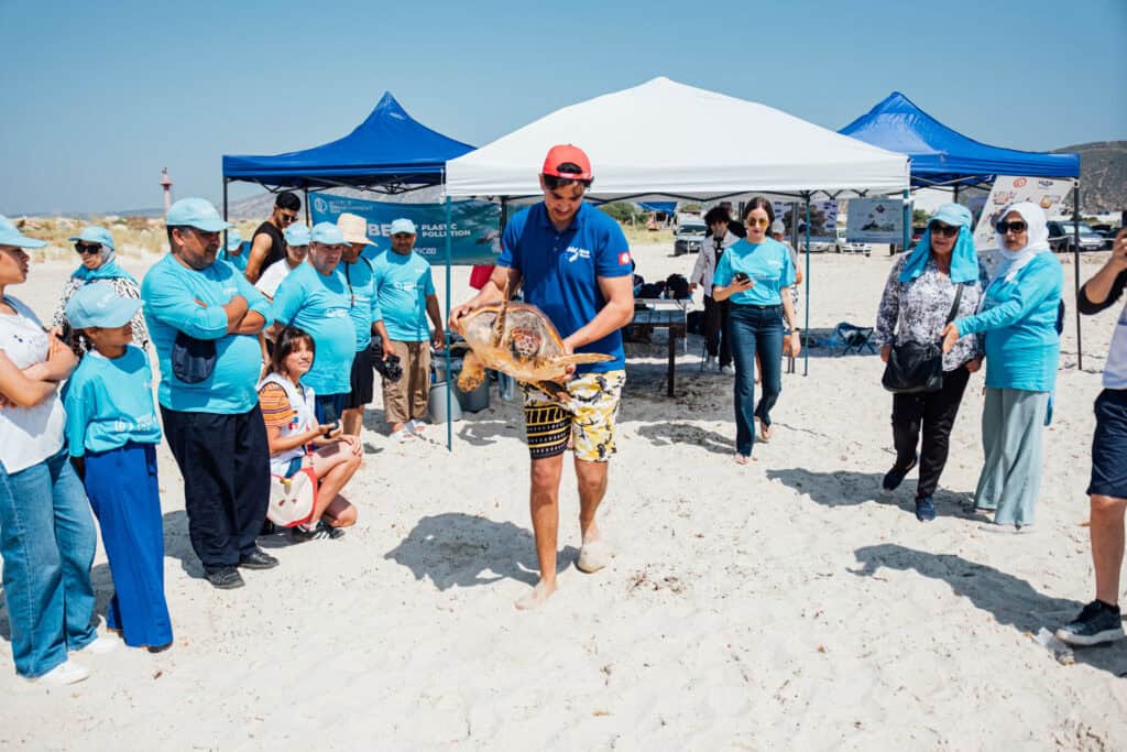 Man in blue shirt carrying sea turtle from sandy beach back toward the ocean in Tunisia